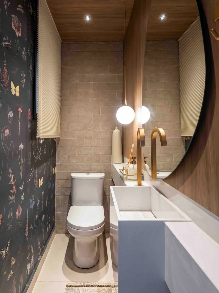 Lavabo com papel de parede floral escuro, metais dourados e espelho redondo